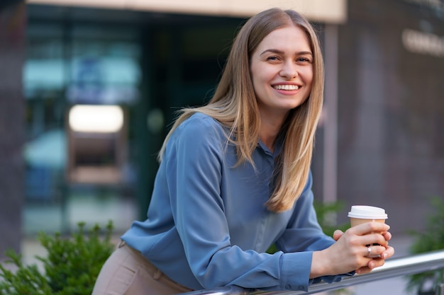 Jonge glimlachende professionele vrouw die een koffiepauze heeft tijdens haar volledige werkdag. Ze houdt een papieren beker buiten bij het bedrijfsgebouw terwijl ze ontspant en geniet van haar drankje.