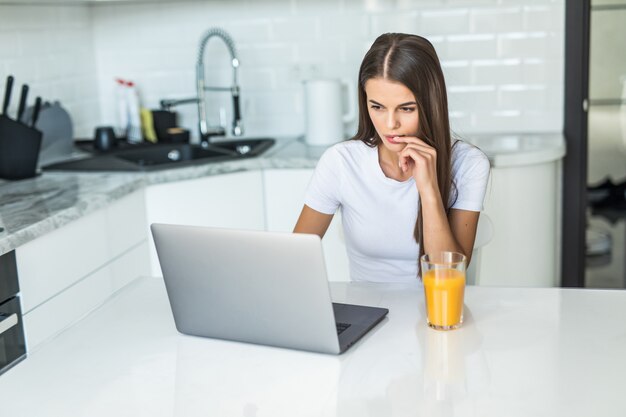 Jonge glimlachende vrouw die ontbijt in de keuken heeft die aan laptop verbindt en een gezond jus d'orange drinkt