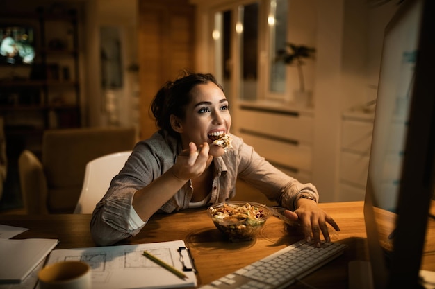 Jonge glimlachende vrouw die op een desktop-pc werkt en 's avonds thuis salade eet