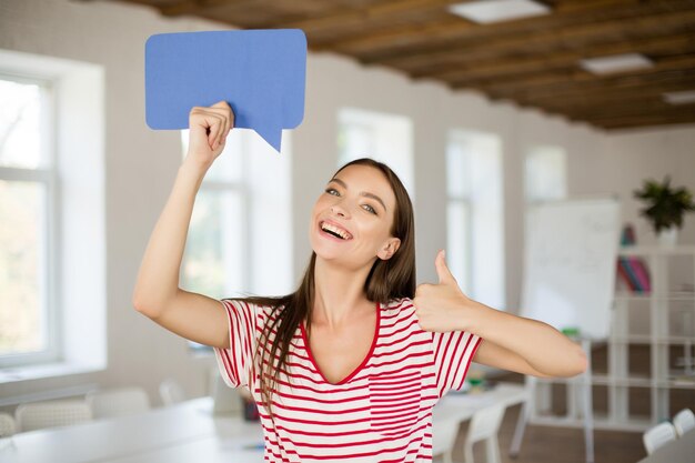 Jonge glimlachende vrouw in gestreept T-shirt die gelukkig in de camera kijkt met het papieren berichtpictogram in de hand in de buurt van het hoofd terwijl ze een duim omhoog gebaar op kantoor toont