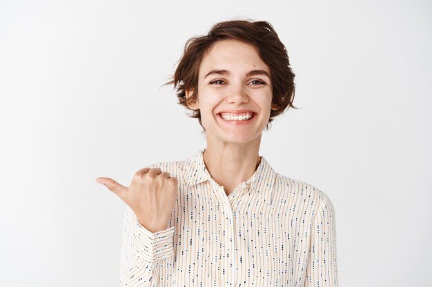 Jonge glimlachende vrouwelijke werknemer die er gelukkig en trots uitziet, met de vinger naar links bedrijf, staande tegen de witte muur