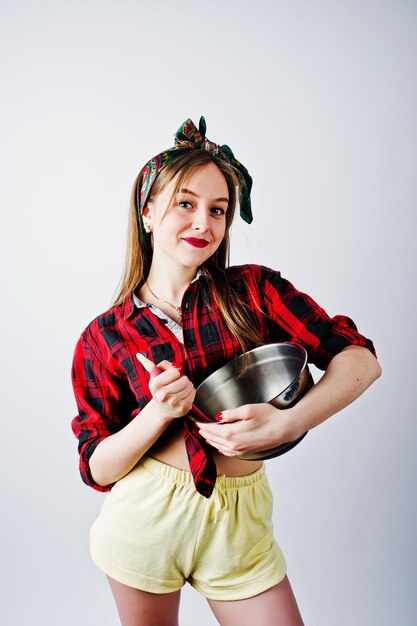 Jonge grappige huisvrouw in geruit overhemd en gele korte broek pin-up stijl met pan en keukenlepel geïsoleerd op een witte achtergrond