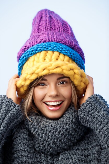 Jonge grappige mooie blonde vrouw in gebreide hoeden en trui lachend op blauw.