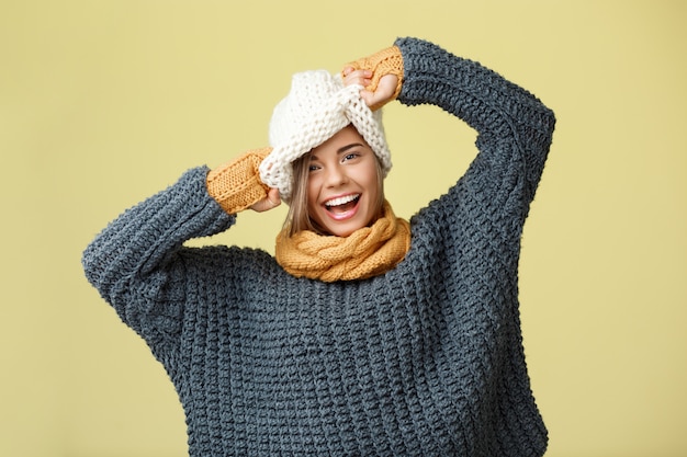 Jonge grappige mooie blonde vrouw in gebreide muts trui sjaal en wanten glimlachend op geel.