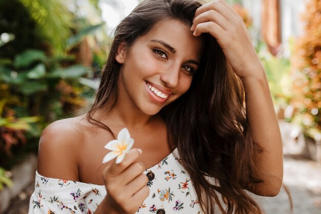 Jonge groenogige dame glimlacht en stelt met witte bloem in tuin