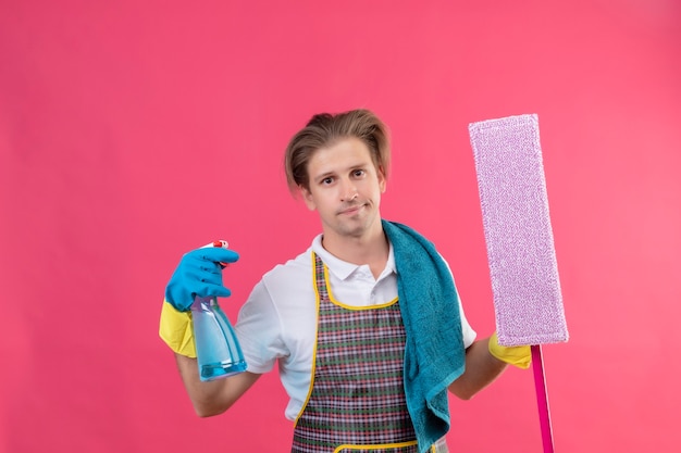 Jonge hansdome-man die schort en rubberhandschoenen draagt die schoonmaaknevel en dweil met sceptische glimlach op gezicht houdt die zich over roze muur bevindt