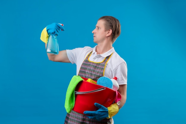 Jonge hansdome-mens die schort en rubberhandschoenen draagt die emmer met schoonmakende hulpmiddelen houden