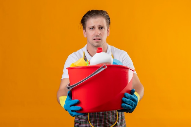 Gratis foto jonge hansdome-mens die schort en rubberhandschoenen draagt die emmer met schoonmakende hulpmiddelen houden