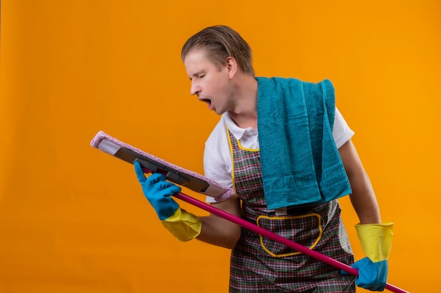 Gratis foto jonge hansdome-mens die schort en rubberhandschoenen draagt die zwabber houdt die het als microfoon gebruikt