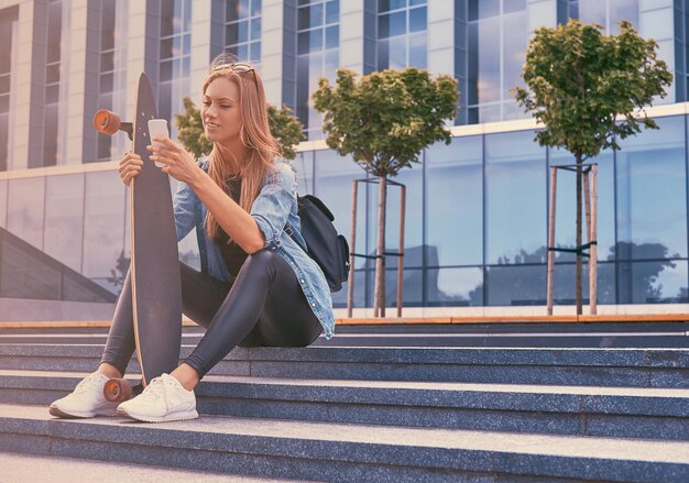 Jonge hipster blonde meisje in casual kleding, zittend op trappen tegen een wolkenkrabber, met behulp van een smartphone, rusten na het rijden op een skateboard.