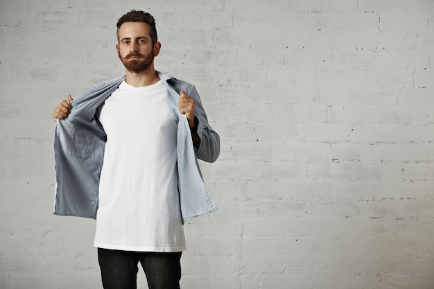 Jonge hipster die zijn verschoten blauwe overhemd met knoopsluiting opstijgt en een wit katoenen t-shirt zonder label op bakstenen muur toont