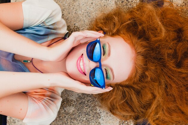 Jonge hipster gember slanke vrouw liggend bij zwembad, uitzicht van bovenaf, rood haar kleurrijk, blauwe zonnebril, sportstijl, sproeten, moedervlekken, ontspannen, gelukkig, speels, coole outfit, glimlachen, sensueel