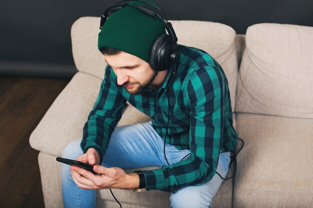 Jonge hipster knappe bebaarde man zittend op een bank thuis, luisteren naar muziek op de koptelefoon