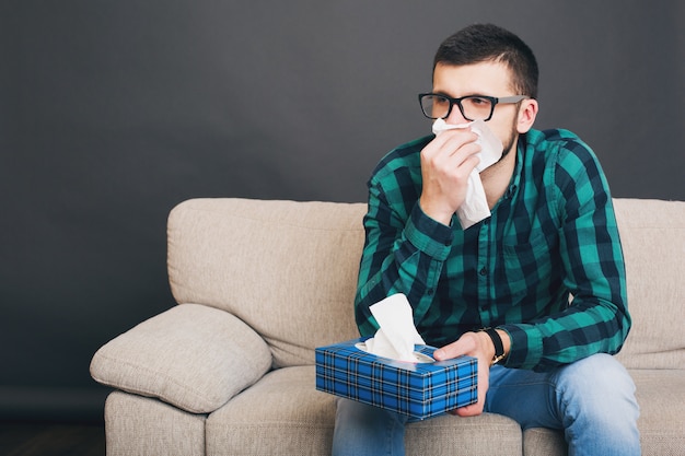 Jonge hipster knappe man in brillen en geruit overhemd om thuis op de bank te zitten, doekjes doos vast te houden, zijn neus in een servet te snuiten, verkouden, ziekte, ziek, griep, overstuur