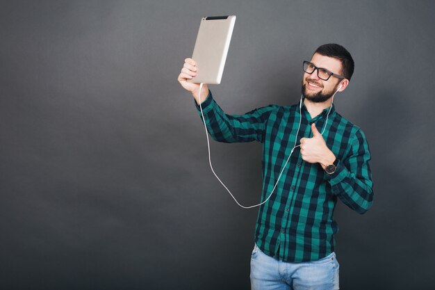 Jonge hipster knappe man op grijze achtergrond houden tablet luisteren naar muziek op oortelefoons praten online gelukkig lachend groen geruit overhemd brillen, positieve stemming