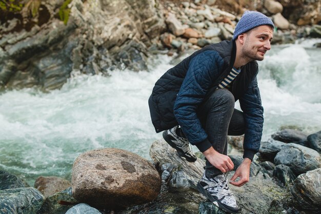 Jonge hipster man lopen op een rots aan de rivier in winter woud, schoenveters koppelverkoop