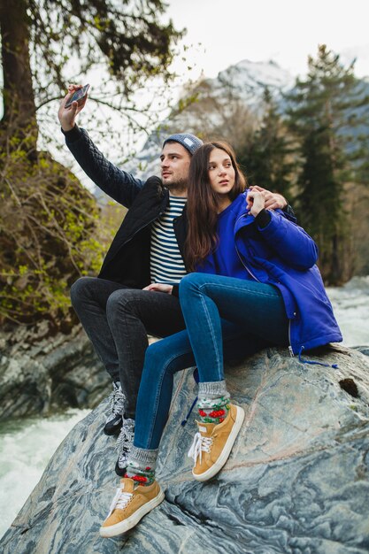 Jonge hipster mooie paar verliefd houden smartphone, fotograferen, zittend op een rots aan de rivier in winter woud