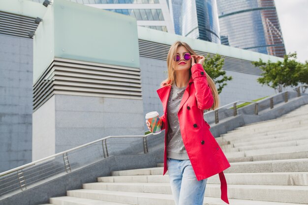 Jonge hipster vrouw in roze jas, jeans in straat met koffie luisteren naar muziek op koptelefoon, zonnebril dragen