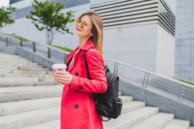 Jonge hipster vrouw in roze jas, jeans in straat met rugzak en koffie luisteren naar muziek op de koptelefoon