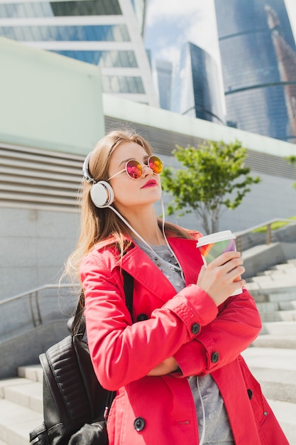 Jonge hipster vrouw in roze jas, jeans in straat met rugzak en koffie luisteren naar muziek op koptelefoon, zonnebril dragen