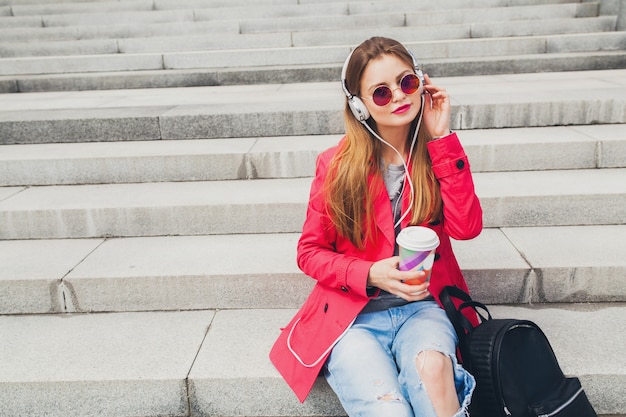 Jonge hipster vrouw in roze jas, jeans in straat met rugzak en koffie luisteren naar muziek op koptelefoon, zonnebril dragen