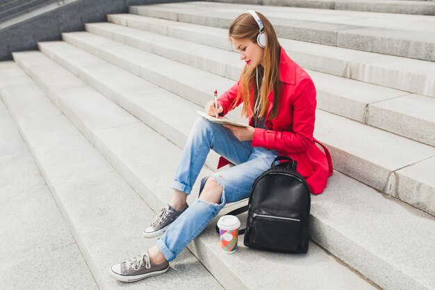 Jonge hipster vrouw in roze jas, jeans zitten in straat met rugzak en koffie luisteren naar muziek op de koptelefoon, student maken van aantekeningen