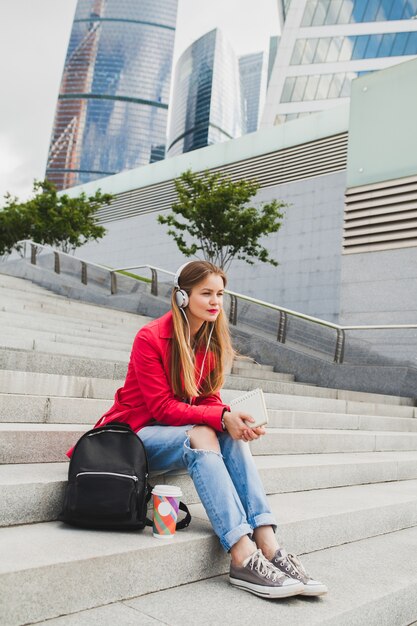 Jonge hipster vrouw in roze jas, jeans zitten in straat met rugzak en koffie luisteren naar muziek op de koptelefoon