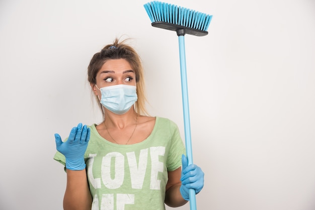 Jonge huisvrouw met gezichtsmasker en bezem die zijwaarts met haar hand op witte muur richt.