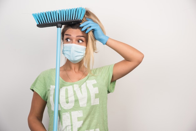 Gratis foto jonge huisvrouw met gezichtsmasker en bezem kijkt verward op witte muur.