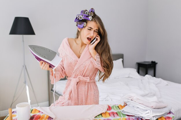 Jonge huisvrouw met lang haar in roze badjas en krultang die ijzer houdt en op telefoon spreekt. Ze ziet er verbaasd uit.