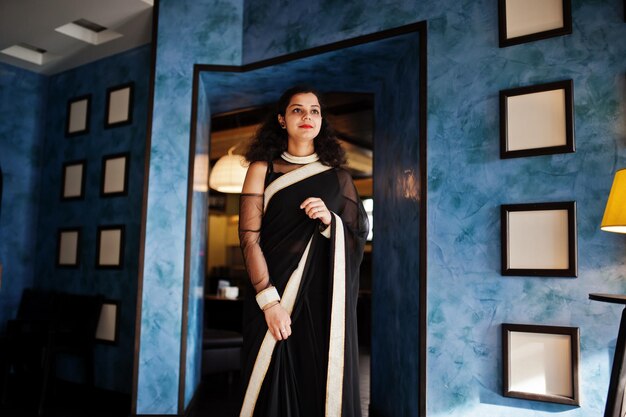 Jonge Indiase vrouw draagt een elegante zwarte sari die op restaurant tegen een muur met frames staat