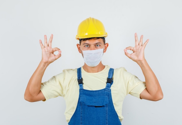 Jonge ingenieur die ok gebaar in uniform, masker vooraanzicht toont.