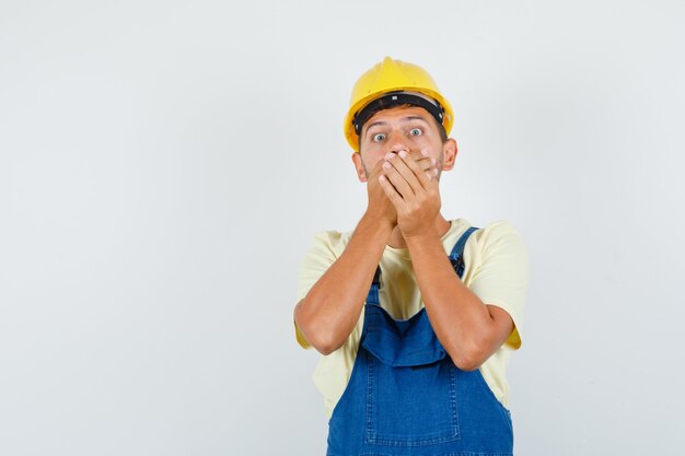 Jonge ingenieur in uniform die mond bedekt met handen en bang, vooraanzicht kijkt.