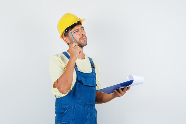 Jonge ingenieur in uniform met potlood en klembord en peinzend, vooraanzicht op zoek.