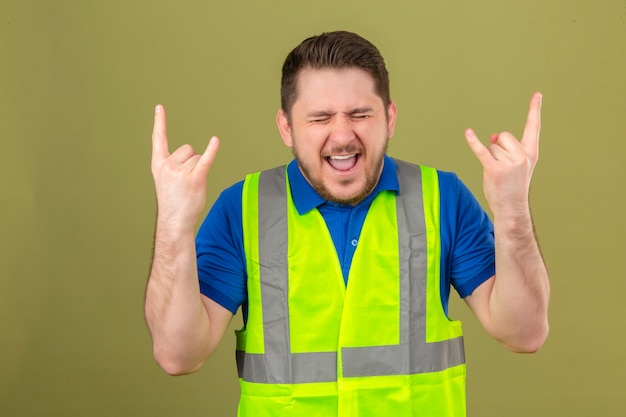 Gratis foto jonge ingenieur man met bouw vest staande met gesloten ogen met gekke uitdrukking doet rock symbool met handen omhoog over geïsoleerde groene achtergrond