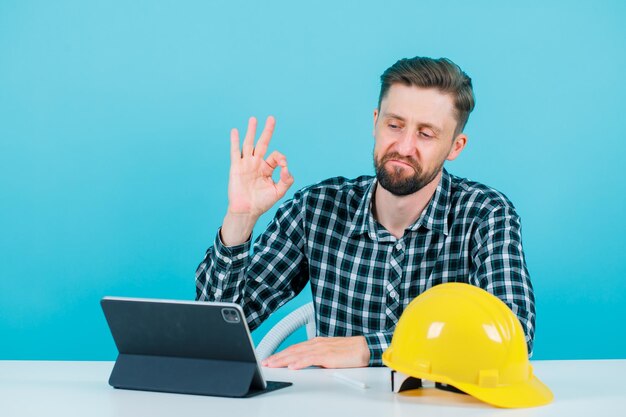 Jonge ingenieur man toont goed gebaar door voor tablet op blauwe achtergrond te zitten