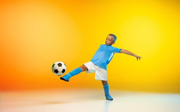 Jonge jongen als een voetbal- of voetbalspeler in sportkleding die oefent op een gele studioachtergrond met gradiënt in neonlicht
