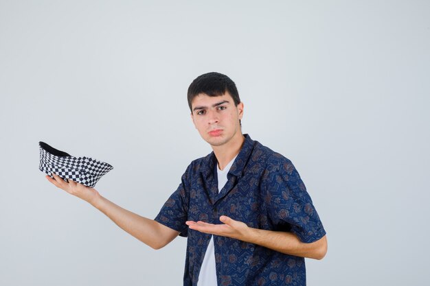 Jonge jongen die de hand uitrekt als een pet in een wit t-shirt, een gebloemd hemd, een pet en er serieus uitziet.