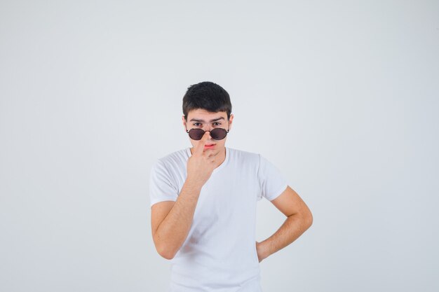 Jonge jongen die de vinger op glazen in t-shirt houdt en koel kijkt. vooraanzicht.