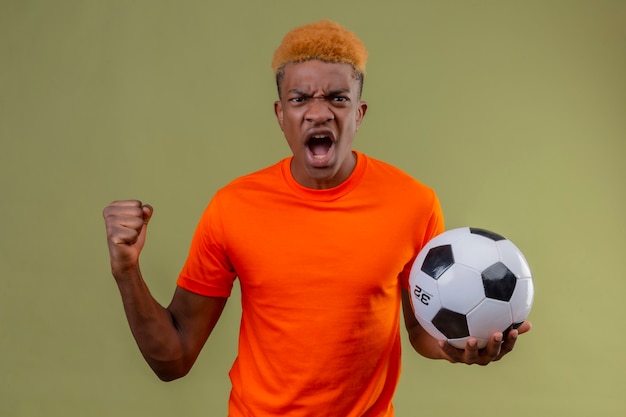 Jonge jongen die oranje t-shirt draagt die de bal balde vuist van de t-shirtholding schreeuwen met boze uitdrukking op gezicht dat zich over groene muur bevindt