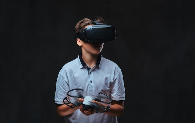 Jonge jongen gekleed in een wit t-shirt met virtual reality-bril en houdt een quadcopter vast. Geïsoleerd op een donkere achtergrond.