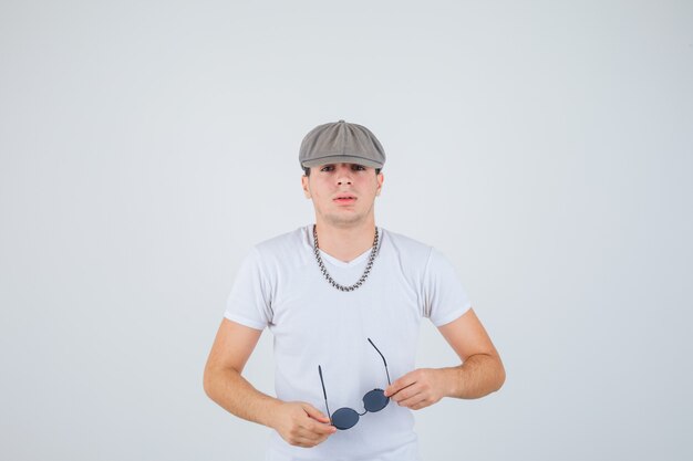 Jonge jongen in t-shirt, hoed met bril, camera kijken en op zoek naar zelfverzekerd, vooraanzicht.