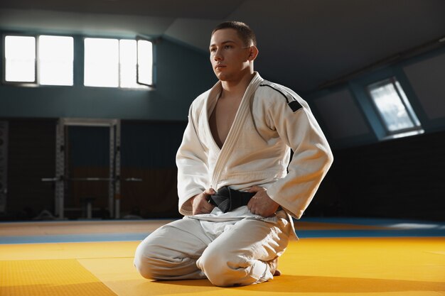 Jonge judo-kaukasische vechter in blauwe kimono met zwarte gordel die zelfverzekerd in de sportschool staat, sterk en gezond.