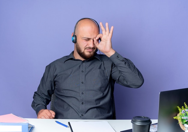 Jonge kale call center man met hoofdtelefoon zittend aan een bureau met uitrustingsstukken doen blik gebaar op laptop geïsoleerd op paarse achtergrond