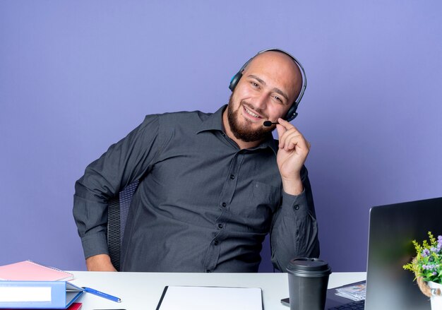 Jonge kale call center man met hoofdtelefoon zittend aan een bureau met uitrustingsstukken geïsoleerd op paarse achtergrond