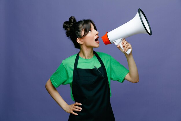 Jonge kappersvrouw die een schort draagt dat in een megafoon spreekt en er zelfverzekerd uitziet over een blauwe achtergrond