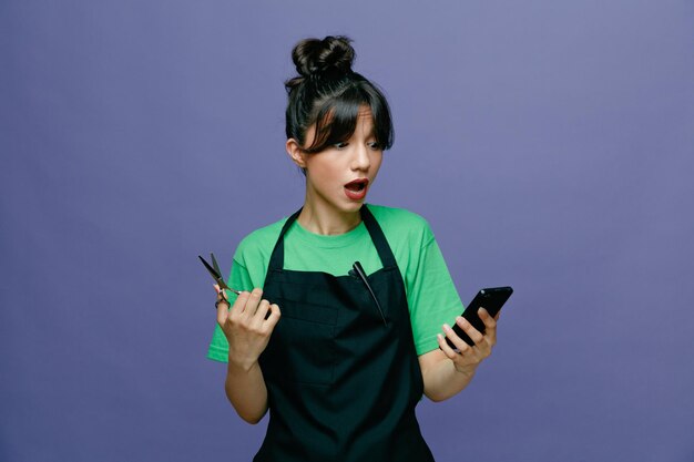 Jonge kappersvrouw die een schort draagt met een mobiele telefoon die een schaar vasthoudt en er verward en verrast uitziet terwijl ze over een blauwe achtergrond staat