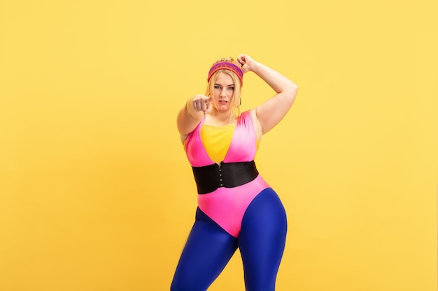 Jonge Kaukasische plus size vrouwelijke model opleiding op gele achtergrond. Kopieerruimte. Concept van sport, gezonde levensstijl, positief lichaam, mode, stijl. Stijlvolle vrouw zelfverzekerd wijzend op.