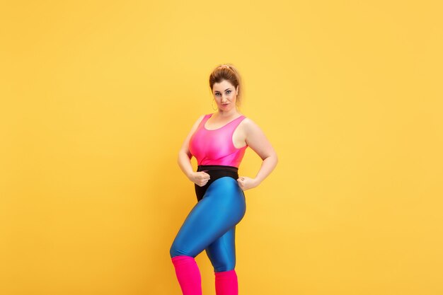 Jonge Kaukasische plus size vrouwelijke model opleiding op gele muur. Kopieerruimte. Concept van sport, gezonde levensstijl, positief lichaam, mode, stijl. Stijlvolle vrouw die zelfverzekerd en cool poseert.