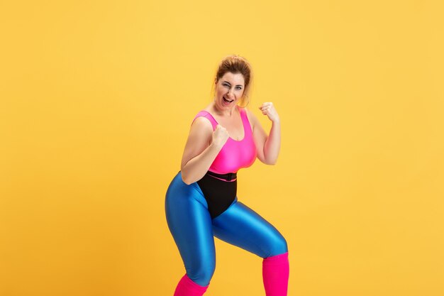 Jonge Kaukasische plus size vrouwelijke model opleiding op gele muur. Kopieerruimte. Concept van sport, gezonde levensstijl, positief lichaam, mode, stijl. Stijlvolle vrouw die zich voordeed als superheld, girl power.